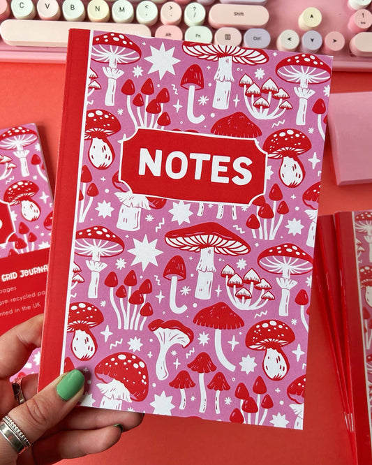 Hand holding the Stacey McEvoy Caunt mushroom notebook, with a full view of the whimsical red and white mushroom design on a pink background. Aesthetic stationery from Rebel Romance, ideal for journaling or drawing.