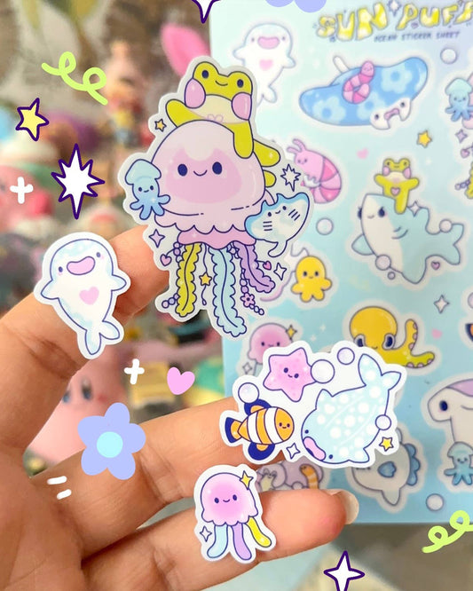 Hand holding several ocean-themed vinyl stickers from Rebel Romance’s Sun Puff Studios Ocean Sticker Sheet. Features kawaii-style characters including a smiling jellyfish with rainbow tentacles, a frog, narwhal, stingray, clownfish and starfish. Bright pastel colours and playful sea creatures ideal for journaling, scrapbooking or gifting.