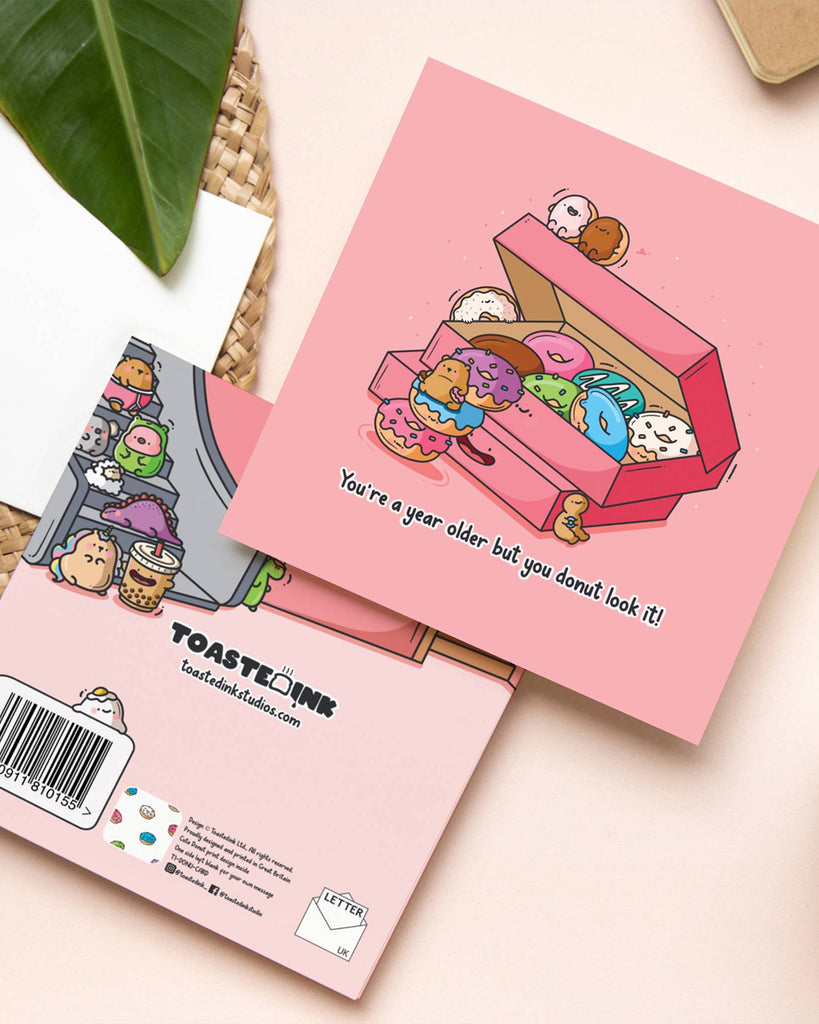 A square pink birthday card by Toastedink featuring a cute box of smiling cartoon donuts and a bear climbing stacked donuts, with the text "You're a year older but you donut look it!" beneath the illustration.
