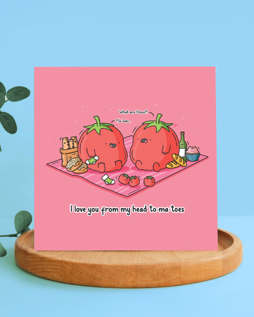 Greeting card standing upright on a wooden plate. Shows two tomatoes on a picnic with snacks, one confused about the other’s “ma toes.” Text below says “I love you from my head to-ma-toes.” A funny and unique Toastedink design, stocked by Rebel Romance.