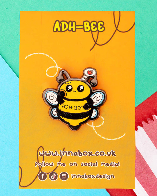 Cute ADH-Bee enamel pin by Innabox on an orange backing card with black and yellow lettering. The pin features a happy yellow bee with glittery black stripes, swirly blue wings, and a heart thought bubble. Sold by Rebel Romance, a UK brand celebrating alternative fashion and millennial style.