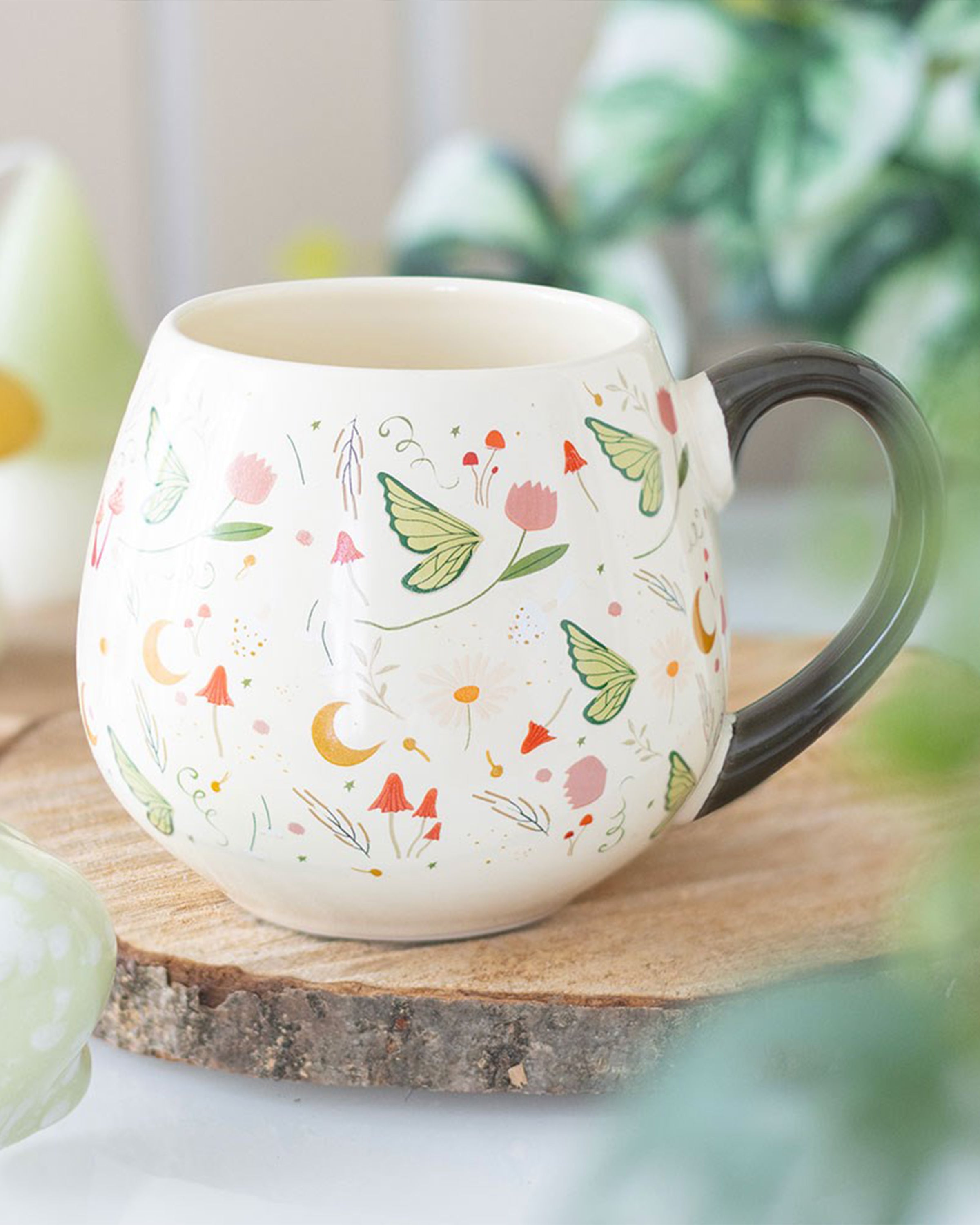 The Rebel Romance Fairy Folklore Print Rounded Mug styled on a wooden coaster with greenery in the background. Its floral, butterfly and moon design creates a cosy magical vibe, making it a unique homeware gift from a UK brand celebrating alternative aesthetics.