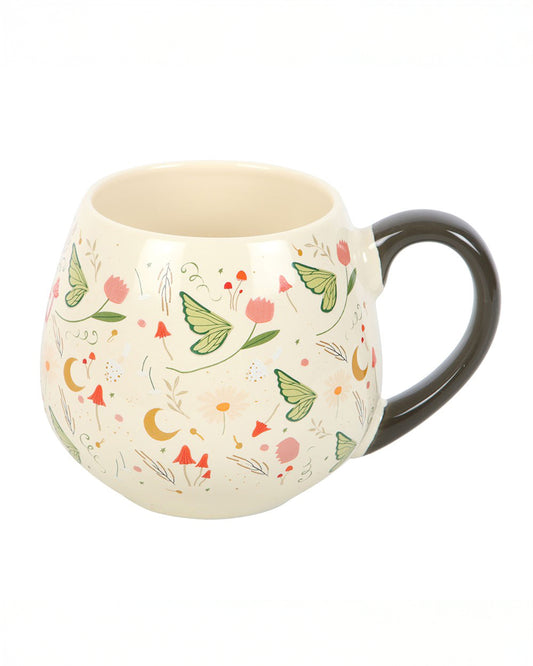 A rounded ceramic mug from Rebel Romance featuring a Fairy Folklore print with green butterflies, pink flowers, mushrooms and golden moons on a cream background. It has a smooth dark handle and is styled for cosy fairycore aesthetics, ideal for warm drinks and magical homeware lovers.