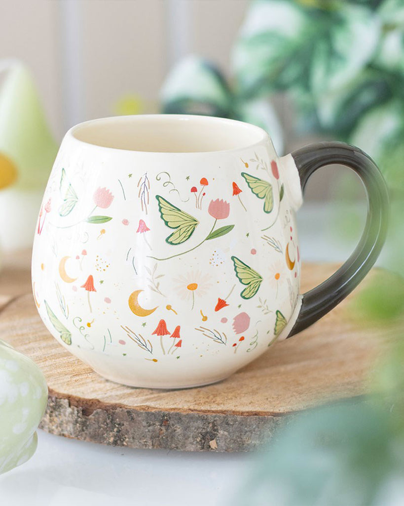 The Rebel Romance Fairy Folklore Print Rounded Mug styled on a wooden coaster with greenery in the background. Its floral, butterfly and moon design creates a cosy magical vibe, making it a unique homeware gift from a UK brand celebrating alternative aesthetics.