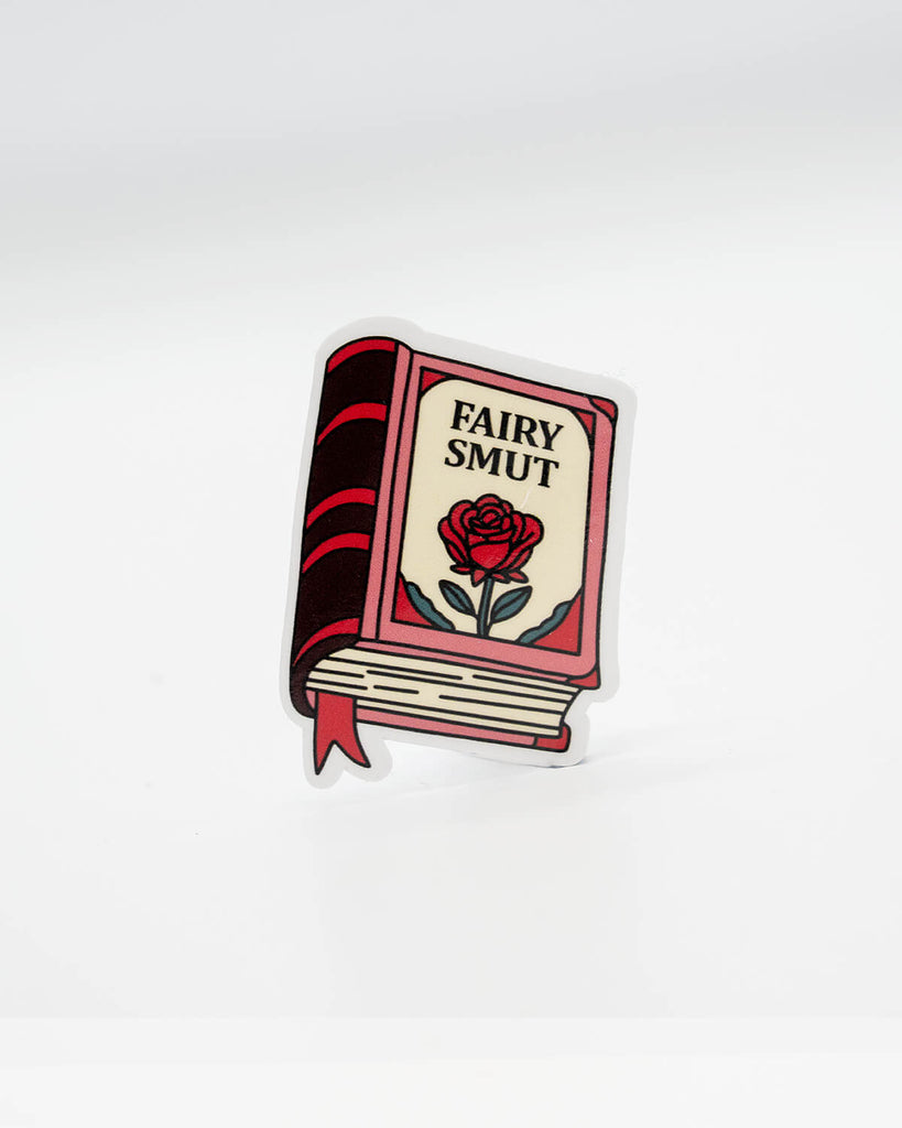 Die-cut vinyl sticker shaped like a red and cream book with the words “Fairy Smut” and an illustrated rose on the cover. A cheeky, bookish decal perfect for lovers of spicy fantasy romance tropes. Designed by Rebel Romance, a small UK brand that celebrates alternative style, slow fashion, and quirky millennial aesthetics through gifts, stickers, and more.