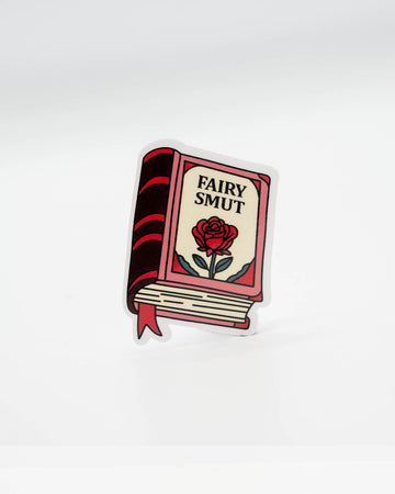 Die-cut vinyl sticker shaped like a red and cream book with the words “Fairy Smut” and an illustrated rose on the cover. A cheeky, bookish decal perfect for lovers of spicy fantasy romance tropes. Designed by Rebel Romance, a small UK brand that celebrates alternative style, slow fashion, and quirky millennial aesthetics through gifts, stickers, and more.