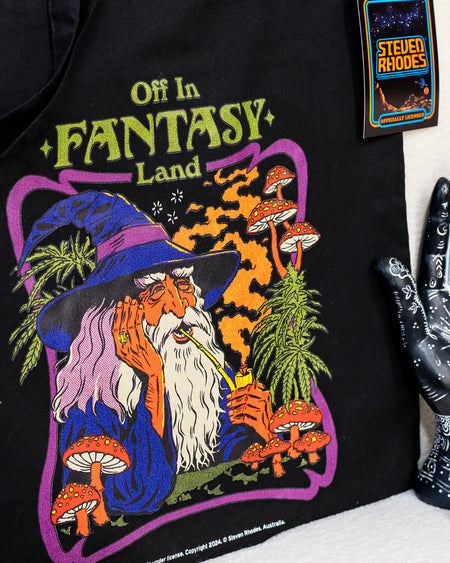 Steven Rhodes black tote bag featuring a whimsical design titled ‘Off in Fantasy Land.’ The artwork depicts a wizard sipping a drink surrounded by mushrooms and magical plants, with bold purple, orange, and green tones. The bag includes sturdy handles and an eco-friendly label, combining functionality with quirky charm.