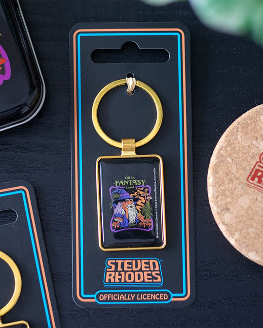 Rebel Romance Steven Rhodes Fantasy Land keyring photographed on a flat lay with other accessories. Gold metal keyring on black Steven Rhodes branded packaging featuring the wizard smoking a pipe surrounded by mushrooms with the phrase “Off in Fantasy Land.” A quirky alternative fashion gift idea.
