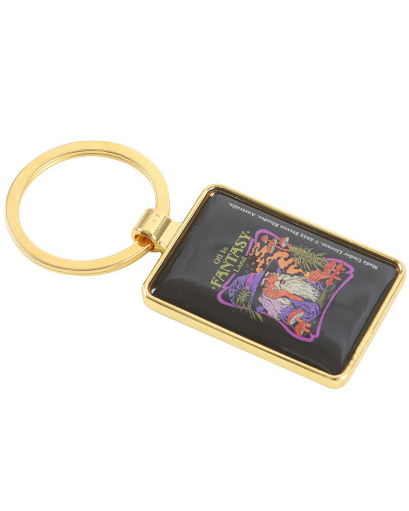 Rebel Romance Steven Rhodes Fantasy Land keyring lying flat. Rectangular gold keyring with a glossy enamel insert showing a wizard in purple robes and hat, smoking a pipe in a trippy mushroom-filled fantasy scene with the words “Off in Fantasy Land.” Perfect gothic and alternative style accessory or gift.