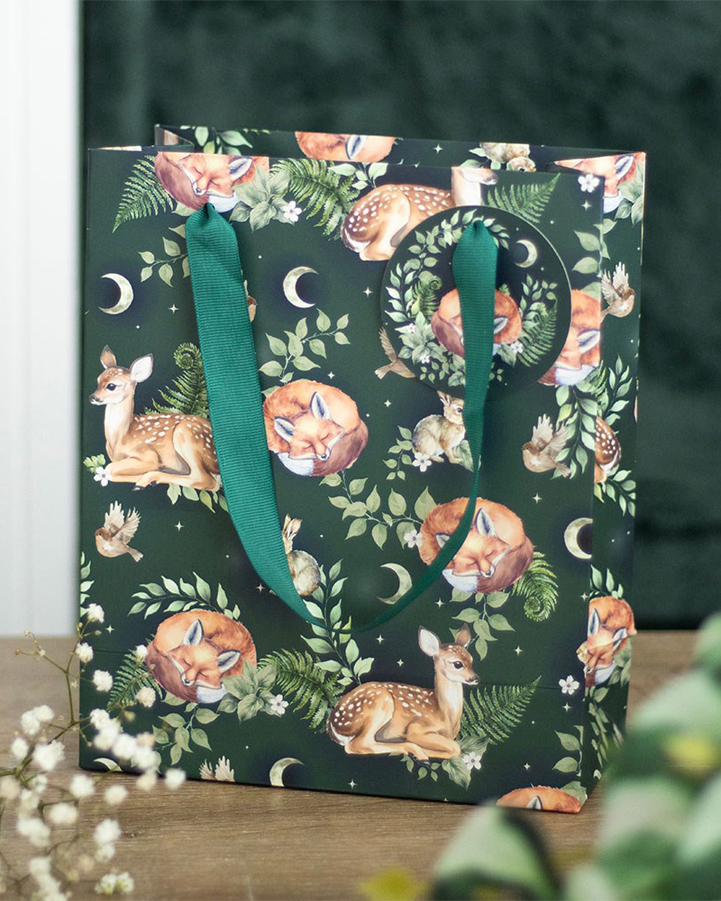 Styled Rebel Romance 23cm Fawn and Friends gift bag on a rustic wooden table, featuring forest animals, ferns and moons on dark green, with matching green ribbon handles and a round gift tag.