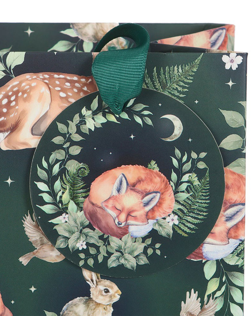 Close-up of a Rebel Romance Fawn and Friends forest gift bag tag, showing a curled fox surrounded by ferns, leaves and stars on a deep green background, attached with a wide green ribbon handle.