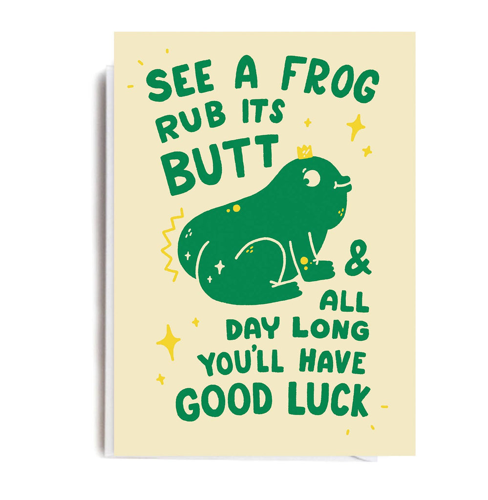 A beige card featuring a green cartoon frog with text that reads “See a frog, rub its butt, & all day long you’ll have good luck,” surrounded by yellow stars and sparkles. Blank inside with a white envelope. Designed by Matt Joyce.