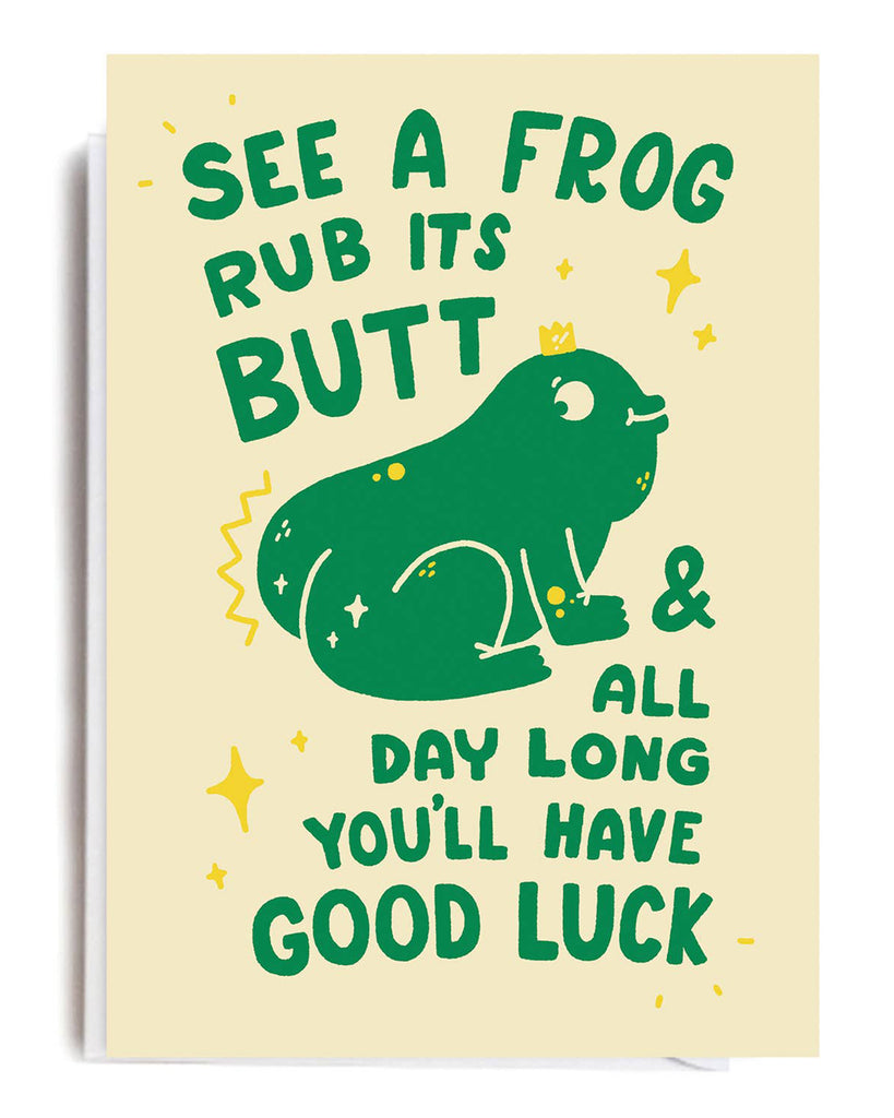 A beige card featuring a green cartoon frog with text that reads “See a frog, rub its butt, & all day long you’ll have good luck,” surrounded by yellow stars and sparkles. Blank inside with a white envelope. Designed by Matt Joyce.