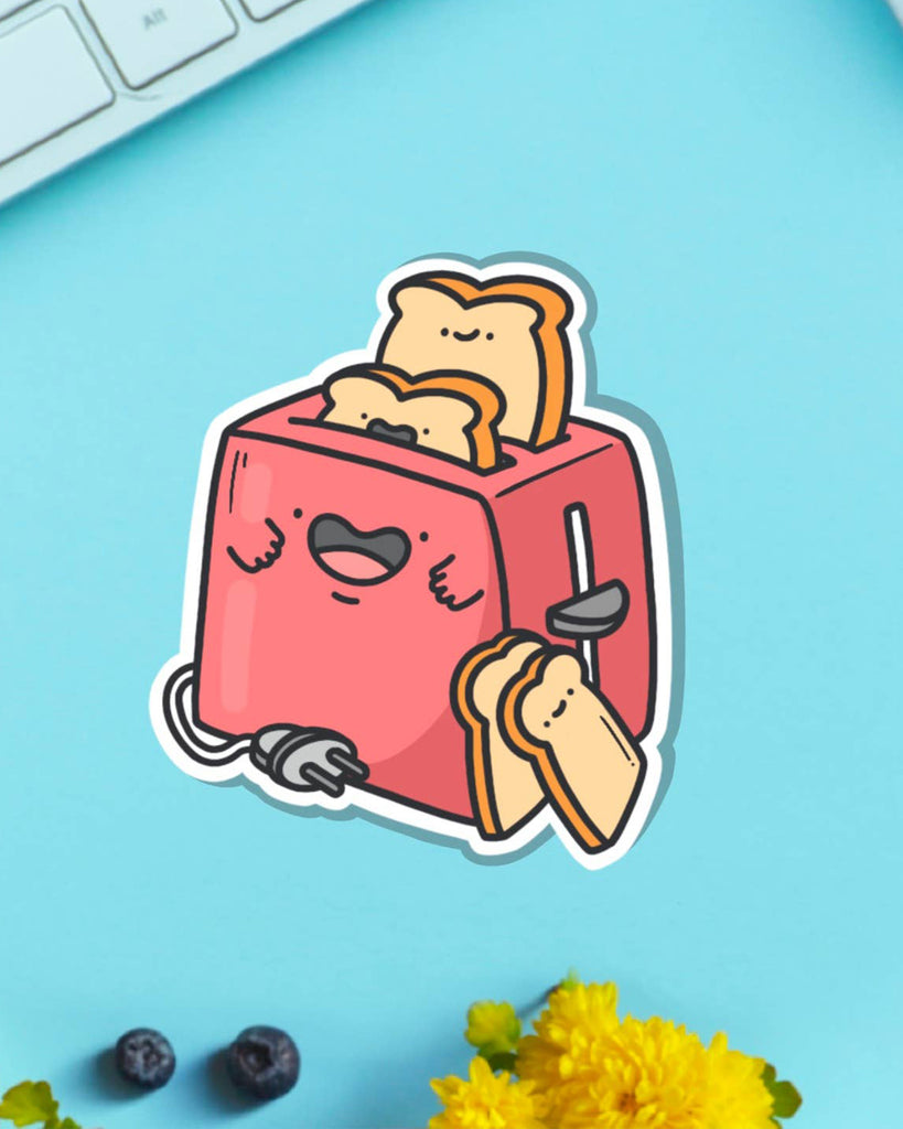 Toastedink’s toaster vinyl sticker on a bright blue desk next to yellow flowers and a white keyboard. Features a red toaster with cute happy toast slices. Sold by Rebel Romance, a UK small biz for alternative, quirky accessories and stationery.