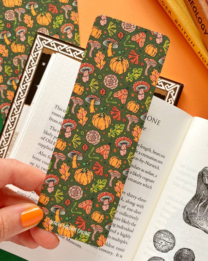 A hand holding an autumnal themed green bookmark filled with hand illustrated autumnal leaves, mushrooms, moths, pumpkins and sparkles. 