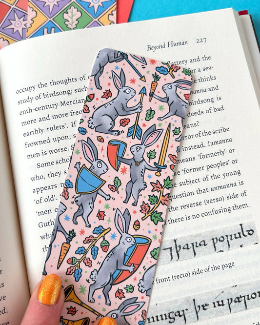 Close-up of the Rebel Romance Medieval Rabbits Bookmark designed by Stacey McEvoy Caunt, placed on an open book. The front features grey rabbits wielding swords and shields on a soft pink background, surrounded by autumn leaves, acorns, and carrots.