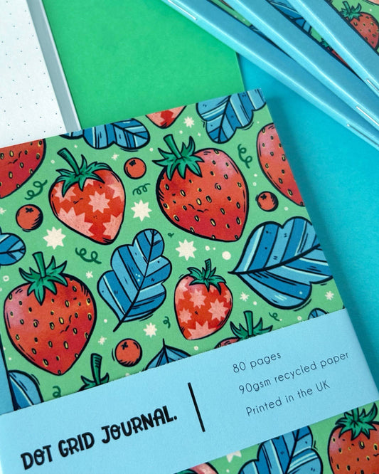 Close-up of the Rebel Romance Strawberry Pocket Notebook with a vivid red strawberry and blue leaf pattern. The notebook is professionally printed with a sky-blue spine and wrap highlighting its eco-friendly recycled paper and UK printing.