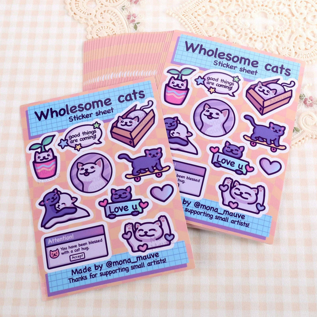 Two Wholesome Cats Sticker Sheets by Mona Mauve laid out on patterned fabric. Each sheet contains 10 adorable vinyl stickers of happy cartoon cats paired with sweet and encouraging messages. Perfect for decorating journals, letters, or gifts. A heartwarming and creative set from a small independent artist.