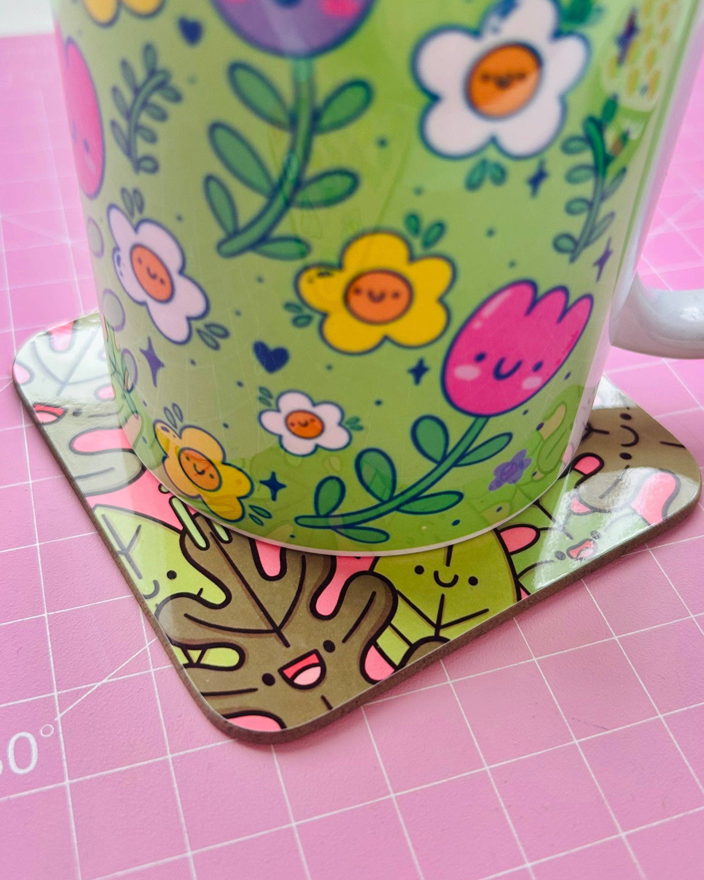 A close‑up of a Rebel Romance drinks coaster by Kelly Lou under a colourful floral mug. The coaster features smiling illustrated leaves on a pink background with a fun, uplifting design. Quirky homeware from this small UK business celebrating alternative style and joyful aesthetics.