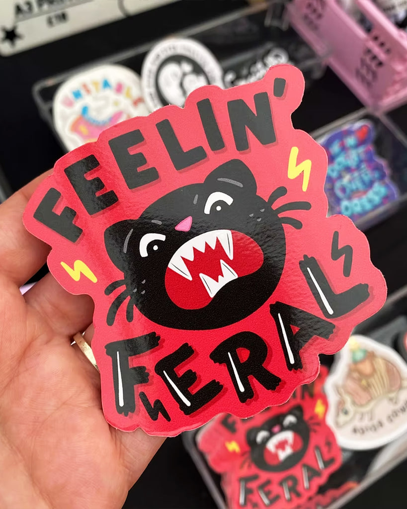 Glossy vinyl sticker from Rebel Romance featuring a fierce black cat illustration with sharp teeth, angry eyes, and pink nose. Surrounded by lightning bolts, the text reads “Feelin’ Feral” in bold black hand lettering on a red background. Designed by Yasmin Cousins. Measures approx. 8cm x 8cm.
