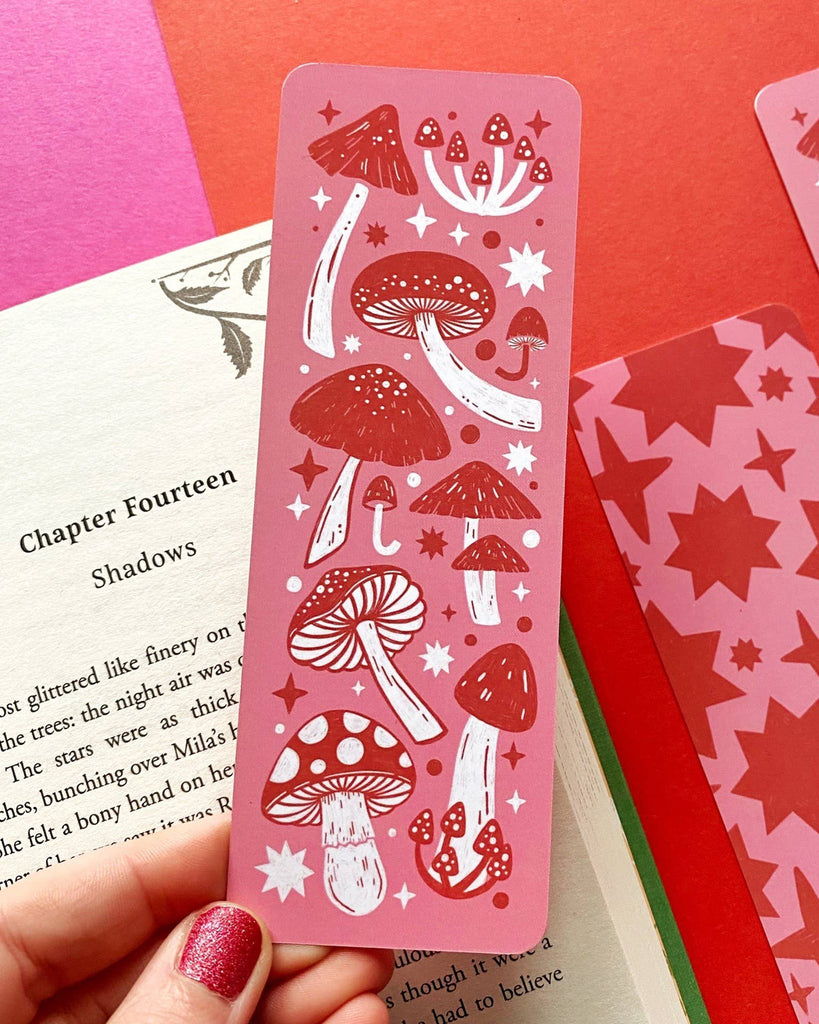 A pink and red mushroom patterned bookmark held between pages of a book, with a person's hand visible at the top with pink nail polish.