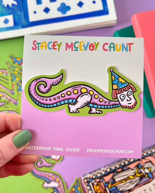 Bright and bizarre vinyl sticker of a medieval-style wizard creature with a human face, pink tail, and pointy hat. Held up against a colourful background with notebooks. A fun, fantasy-inspired waterproof sticker by Rebel Romance.