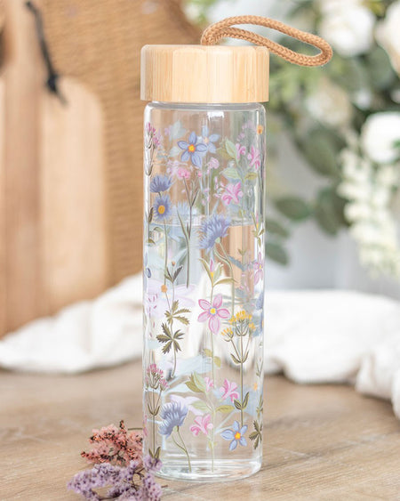Rebel Romance Floral Print Glass Water Bottle styled on a wooden surface with flowers and greenery. The pastel wildflower design pops against the clear glass. Featuring a bamboo lid with a rope handle, it's a perfect blend of beauty and practicality.