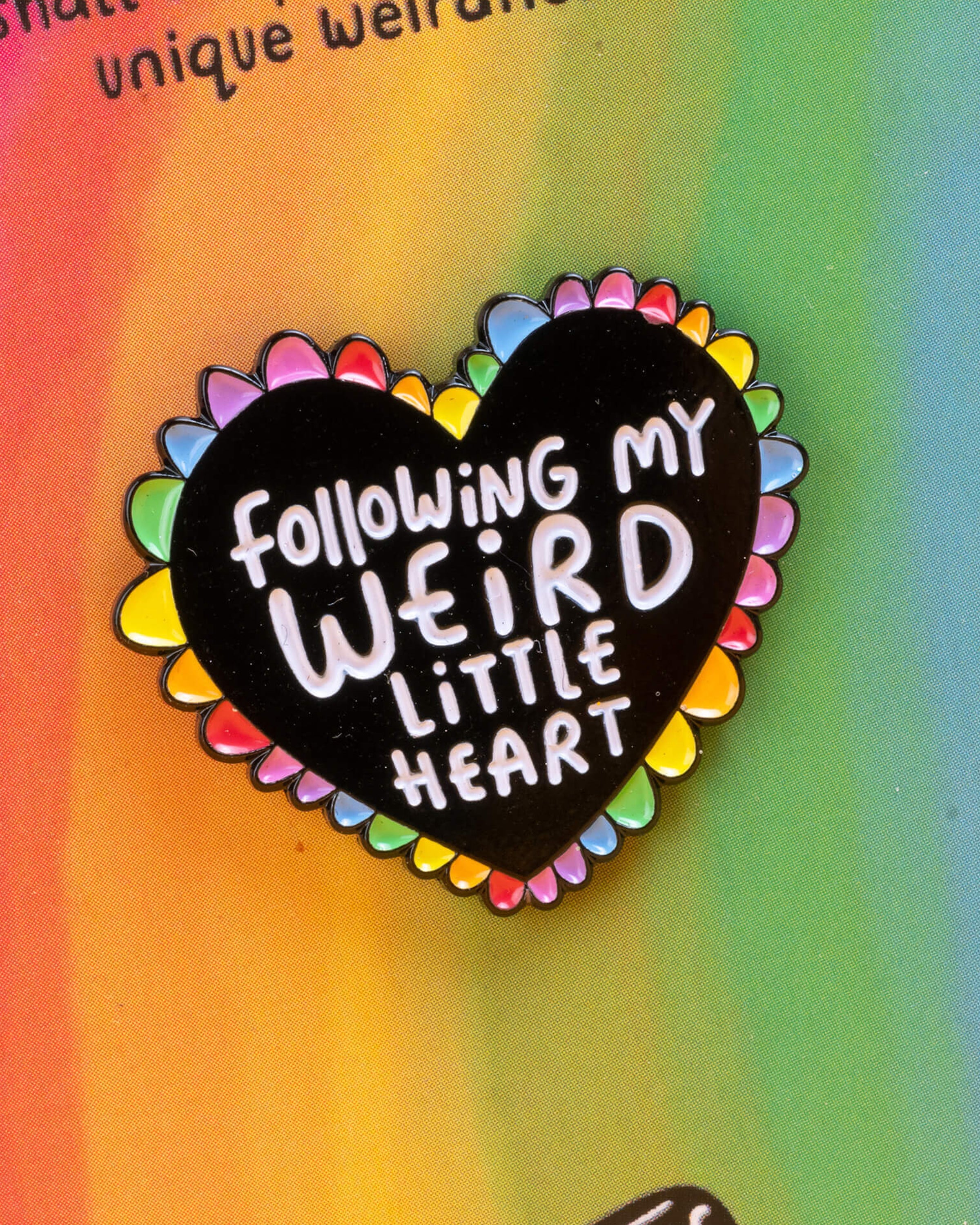 Close-up of a Rebel Romance Weird Little Heart enamel pin badge on a rainbow background card. The black heart-shaped pin has a rainbow edge and the words “Following My Weird Little Heart” in white lettering, celebrating individuality and alternative style.