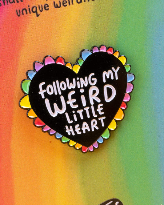 Close-up of a Rebel Romance Weird Little Heart enamel pin badge on a rainbow background card. The black heart-shaped pin has a rainbow edge and the words “Following My Weird Little Heart” in white lettering, celebrating individuality and alternative style.
