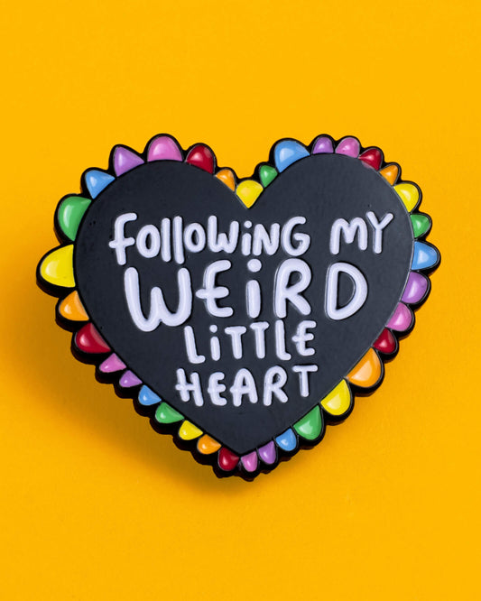 Rebel Romance Weird Little Heart enamel pin badge on a bright yellow background. The black heart-shaped pin features a colourful rainbow edge and white lettering reading “Following My Weird Little Heart,” perfect for alternative fashion and self-expression.