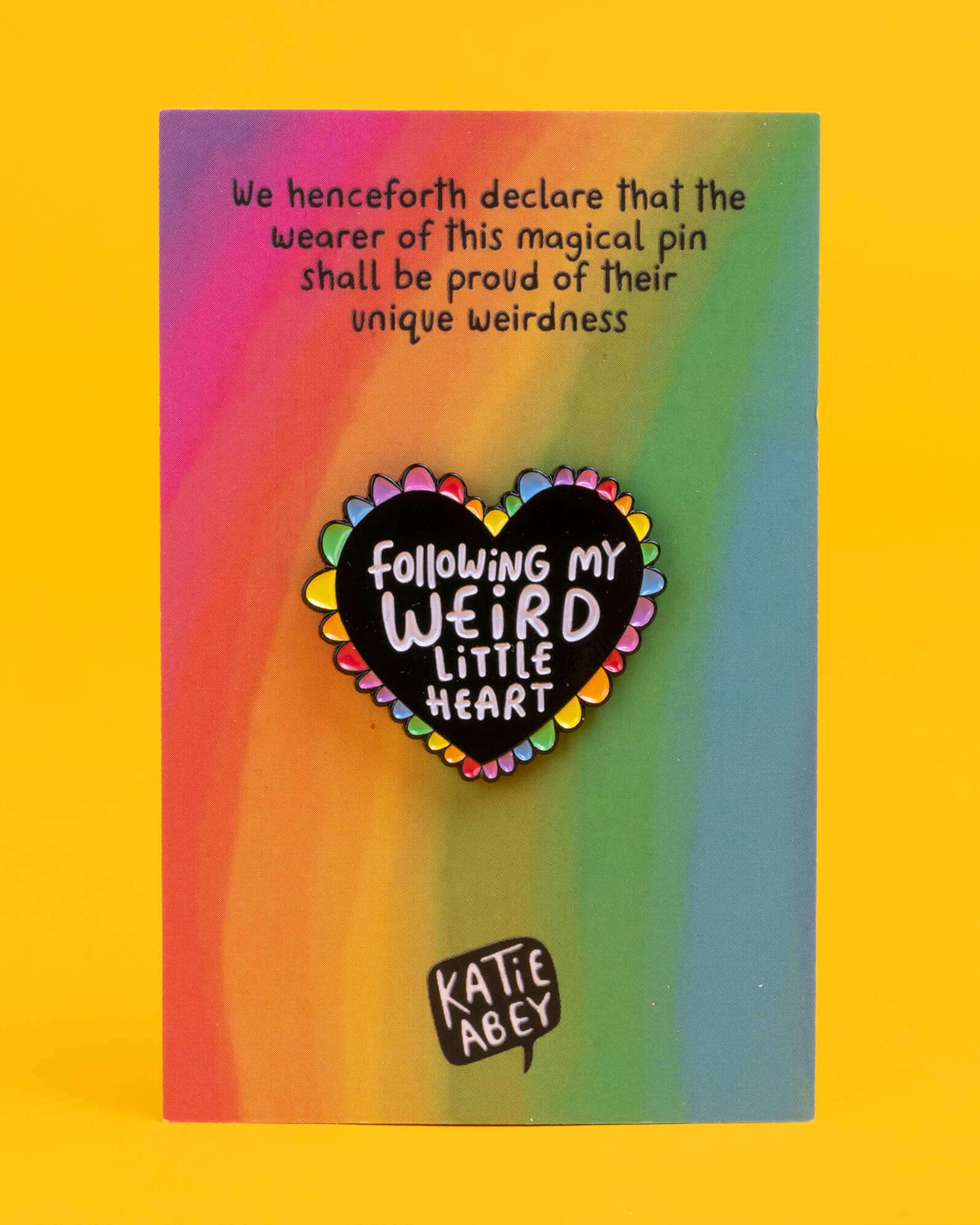 Rebel Romance Weird Little Heart enamel pin badge displayed on its backing card with a rainbow gradient and playful text, ideal for alternative fashion lovers. The pin is a black heart with rainbow edging and white lettering reading “Following My Weird Little Heart.”