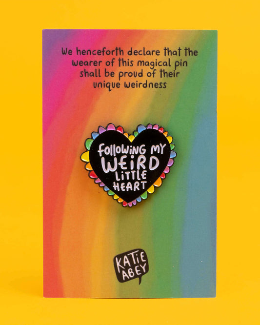 Rebel Romance Weird Little Heart enamel pin badge displayed on its backing card with a rainbow gradient and playful text, ideal for alternative fashion lovers. The pin is a black heart with rainbow edging and white lettering reading “Following My Weird Little Heart.”