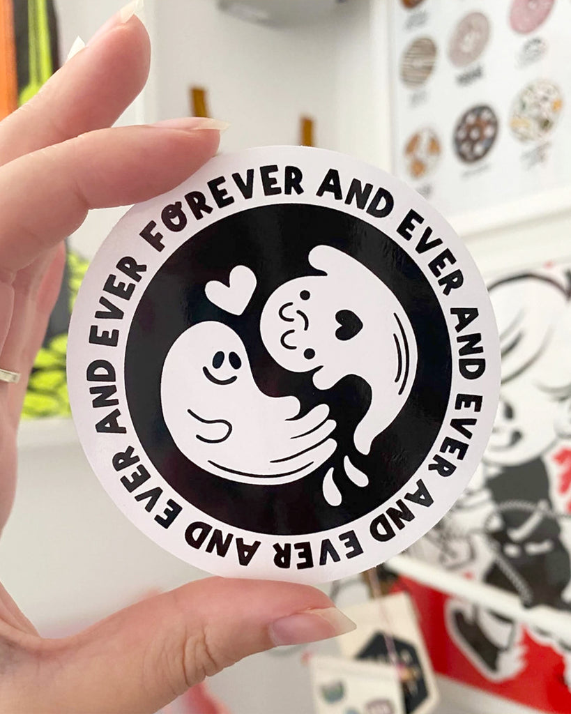 A hand holds up a round black-and-white vinyl sticker stocked by Rebel Romance, featuring two ghost characters with hearts between them, smiling sweetly. The design reads “FOREVER AND EVER AND EVER AND EVER” in a circle around the edge. A quirky and romantic sticker designed by Yasmin Cousins, perfect for alternative style lovers.