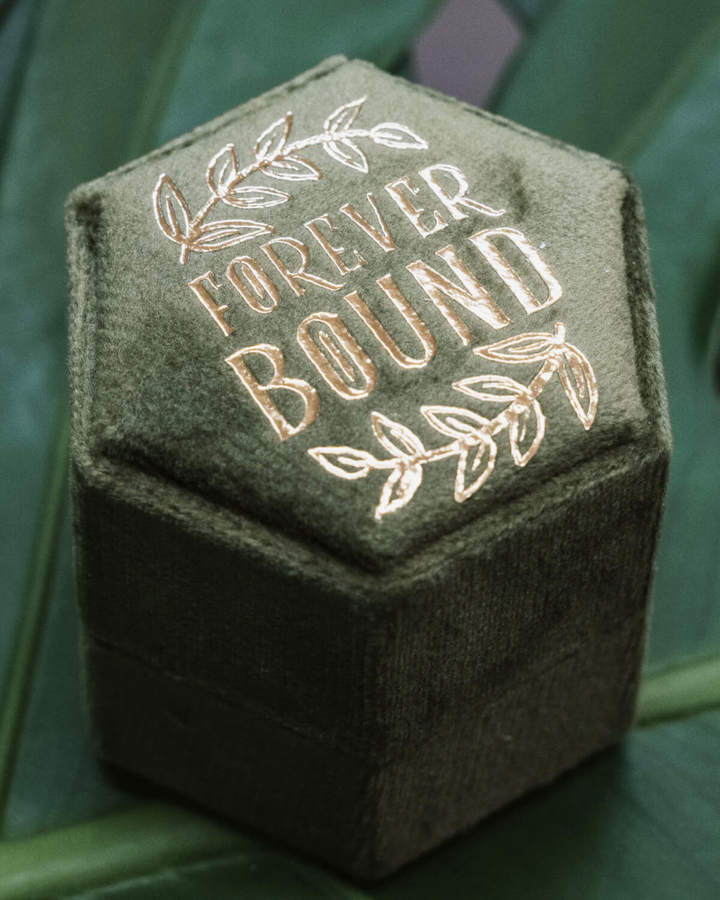 A close-up of the “Forever Bound” hexagonal ring box in dark green velvet with the words “Forever Bound” embossed in shiny gold foil and framed by delicate leaf details. The box is displayed upright on a green monstera leaf.
