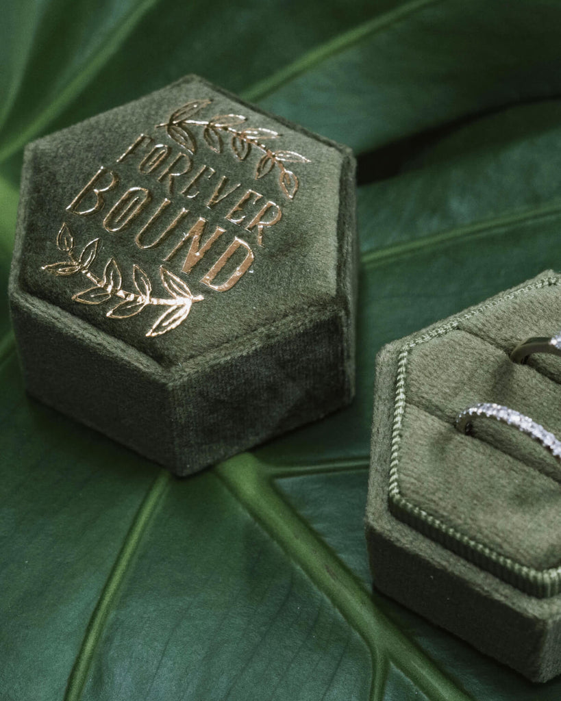 Two halves of the “Forever Bound” hexagonal velvet ring box displayed open on a green monstera leaf, showcasing two rings inside—a solitaire engagement ring and a matching diamond band. The box is deep green with gold foil text and leaf accents.