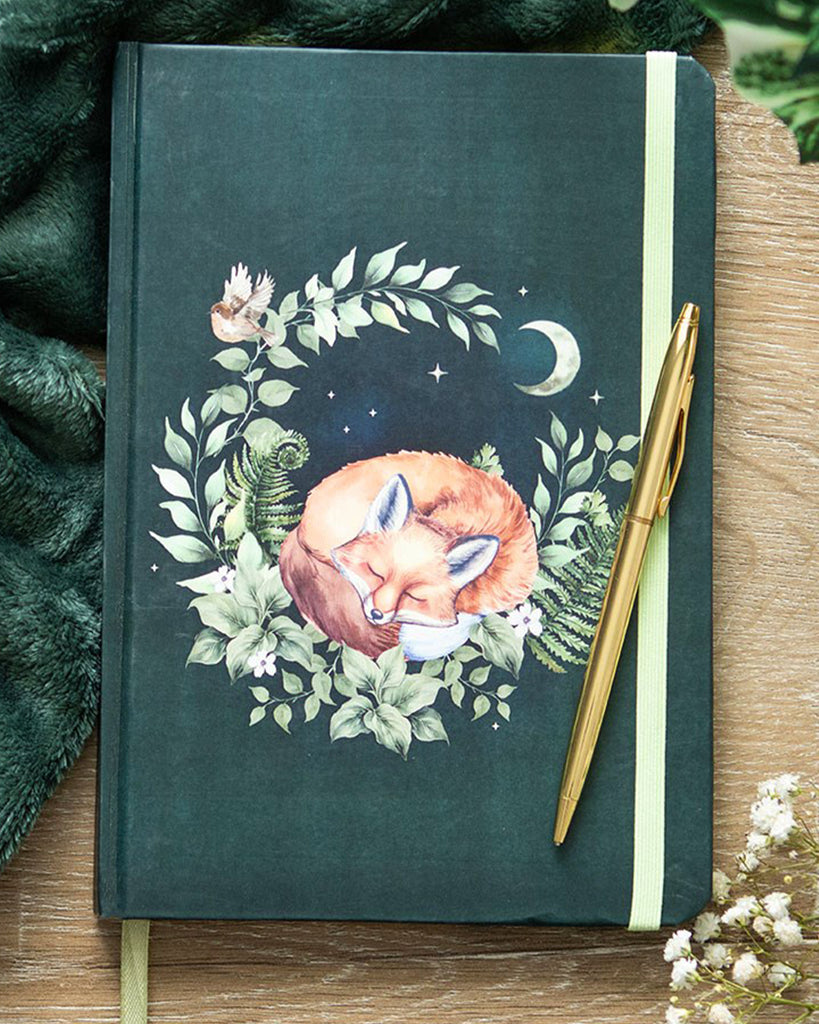 Green Sleeping Fox A5 Notebook
Alt text: An A5 notebook with a forest green cover featuring a curled-up fox sleeping within a circle of leaves, flowers, and a crescent moon. Surrounded by cosy decor and a gold pen.