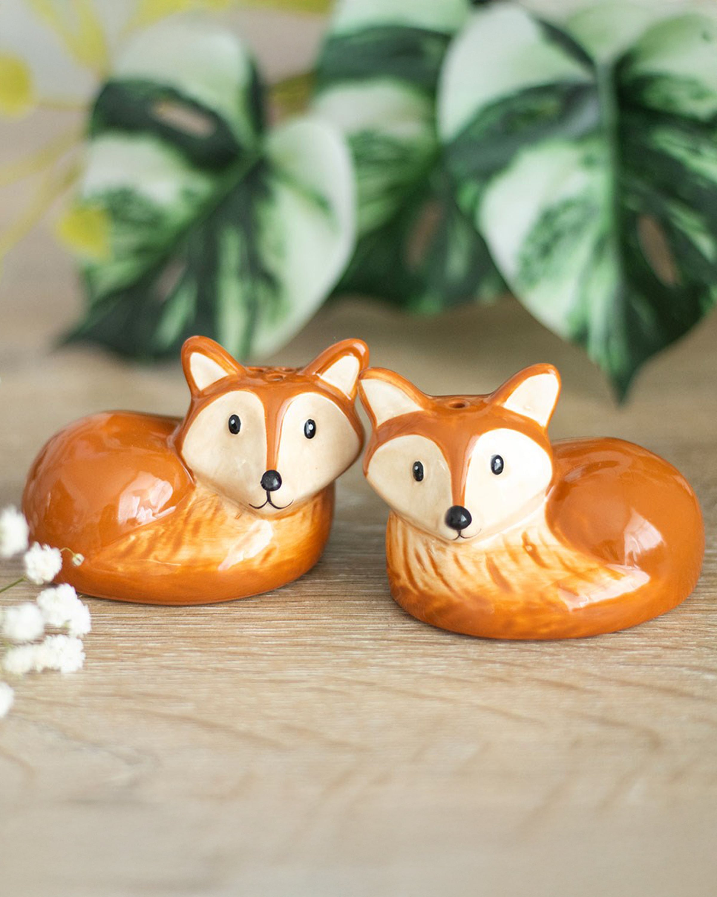 A pair of ceramic salt and pepper shakers shaped like curled-up foxes, with orange bodies, cream faces, and black eyes and noses, displayed on a wooden surface with leafy background.