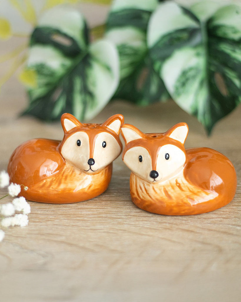 A pair of ceramic salt and pepper shakers shaped like curled-up foxes, with orange bodies, cream faces, and black eyes and noses, displayed on a wooden surface with leafy background.