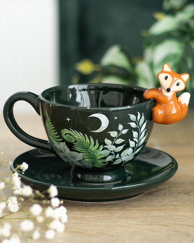 Rebel Romance green forest teacup with orange fox and foliage design shown on a rustic wooden surface with flowers in the foreground. A perfect cottagecore gift for tea lovers.