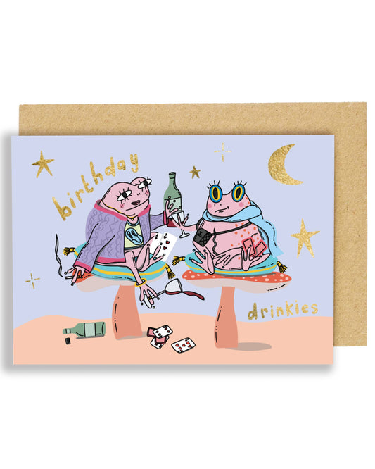 Illustrated birthday card showing two quirky frogs sitting on colourful mushrooms, holding drinks and playing cards under gold foil stars and a crescent moon, with “birthday drinkies” in gold foil, paired with a brown Kraft envelope.