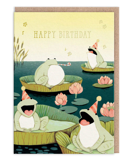 A6 birthday card featuring cute frogs wearing party hats and singing on lily pads in a pond, surrounded by lotus flowers. Designed by Joo Joo Paper and stocked by Rebel Romance, this card includes neon coral details and a kraft envelope. A whimsical, nature-themed birthday card perfect for frog lovers, musicians, and fans of joyful alternative stationery.