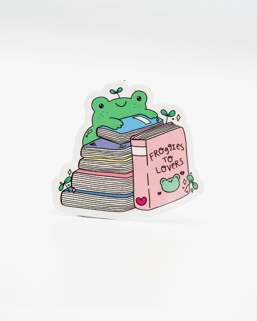 Cute die-cut vinyl sticker featuring a smiling green frog lounging on a stack of pastel books, with the largest book titled “Froggies to Lovers.” Surrounded by hearts, sparkles, and leafy sprouts, this playful design is perfect for frog fans and romance readers. Created by Rebel Romance, a small UK brand celebrating alternative style, slow fashion, and quirky, millennial-friendly bookish gifts.