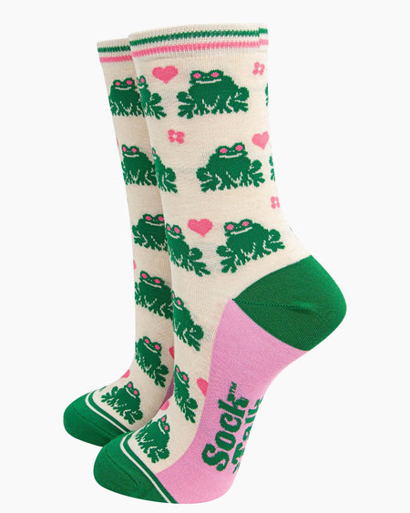 A pair of cream-coloured bamboo socks featuring a fun green frog pattern with pink love hearts and flowers. The socks have a striped green and pink cuff, green heels, and pink soles with ‘Sock Talk’ branding