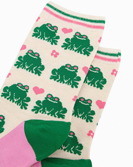 A pair of cream-coloured bamboo socks featuring a fun green frog pattern with pink love hearts and flowers. The socks have a striped green and pink cuff, green heels, and pink soles with ‘Sock Talk’ branding