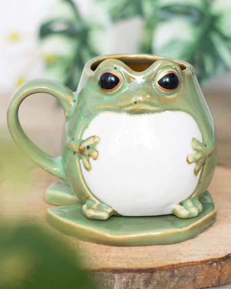 The frog mug placed on a wooden surface with a blurred green plant in the background. The unique design makes it a charming gift for frog lovers and fans of quirky homeware.