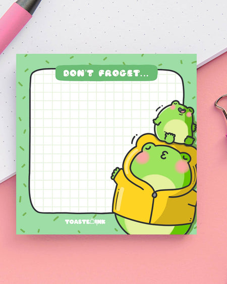A square frog-themed sticky note pad with a green grid background, featuring a chubby frog in a yellow raincoat being kissed by a smaller frog. The top reads “Don’t Froget…” in a white font on a green tab. Perfect for cute reminders and dopamine-fuelled planning.