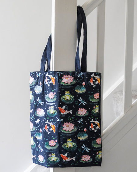 A navy blue polycotton tote bag with colourful pond life illustrations, including frogs, koi fish, lily pads, and dragonflies, hanging from a white stair banister. The bag has long handles and a black back panel.