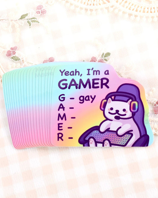 Stack of Rebel Romance vinyl stickers by Mona Mauve featuring a white cartoon cat in a gaming chair with rainbow headset and keyboard, alongside the text “Yeah, I’m a GAMER – G for gay.” Printed on a pastel rainbow gradient background, this waterproof and durable sticker is ideal for laptops, consoles, and journals. A fun and affirming pride-themed design for queer gamers and millennials who love cute, alternative stationery with humour.