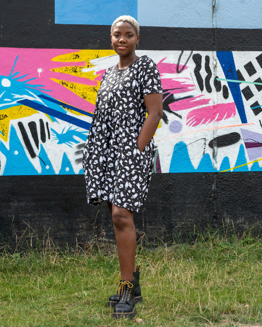 Chish models a stretch cotton smock dress featuring an all-over white ghost print. This alternative casual dress has short sleeves, side pockets, and a relaxed fit, ideal for everyday gothic and quirky style.