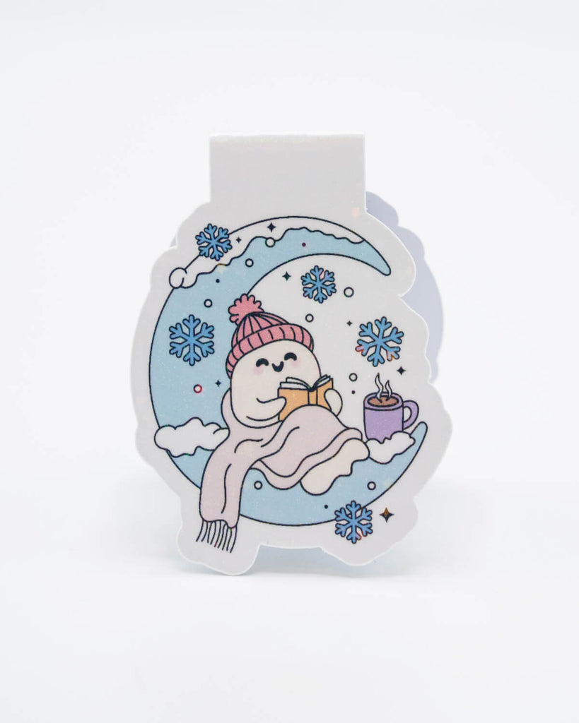Front view of the Winter Reads Ghost Magnetic Bookmark from Rebel Romance, showing a smiling ghost in a pink hat and scarf reading a book on a snowy crescent moon. With pastel snowflakes, clouds, and a mug of hot chocolate, it’s the perfect accessory for fans of cosy winter aesthetics and spooky-sweet style.